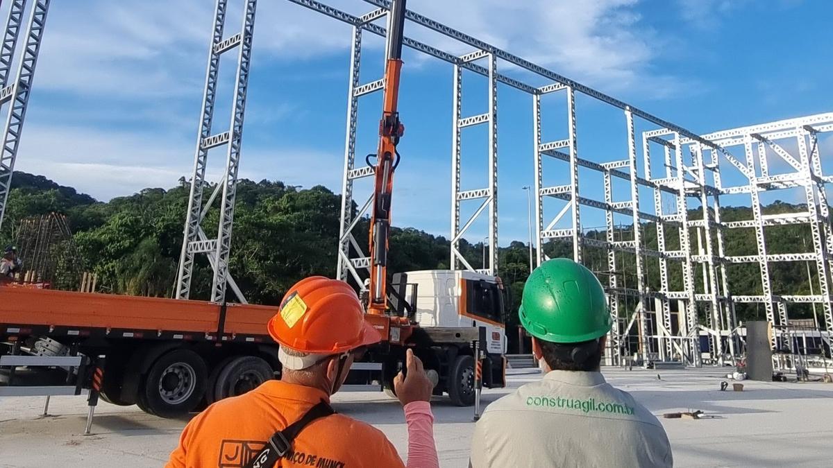 Dois técnicos com capacete acompanham a montagem de uma estrutura em light steel
              frame, com a grua a posicionar painéis ao fundo.